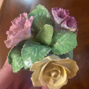 May include: A porcelain figurine of a bouquet of flowers. The bouquet includes a yellow rose, a pink carnation, and a purple carnation. The flowers are surrounded by green leaves.