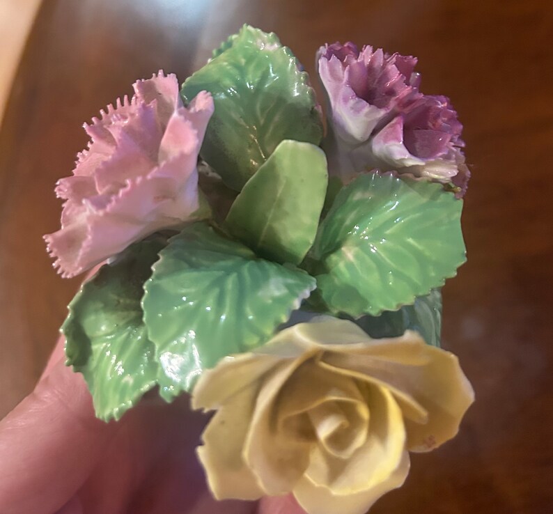 May include: A porcelain figurine of a bouquet of flowers. The bouquet includes a yellow rose, a pink carnation, and a purple carnation. The flowers are surrounded by green leaves.