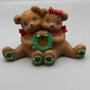 May include: A brown ceramic figurine of two teddy bears embracing. Each bear has a red bow and a green and gold wreath. The bears are sitting and have green and red accents on their feet.