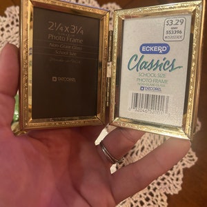 May include: A gold-colored, hinged photo frame with a black mat and non-glare glass. The frame is labeled "2 1/4 x 3 1/4" and "School Size". The frame is made in the USA and is a product of "DECOREL".