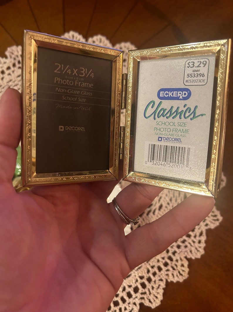 May include: A gold-colored, hinged photo frame with a black mat and non-glare glass. The frame is labeled "2 1/4 x 3 1/4" and "School Size". The frame is made in the USA and is a product of "DECOREL".