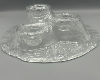 May include: Clear glass candle holder with three circular wells and a wide, textured base. The glass has a crackled appearance, adding a unique texture to the piece. A decorative item for home decor.