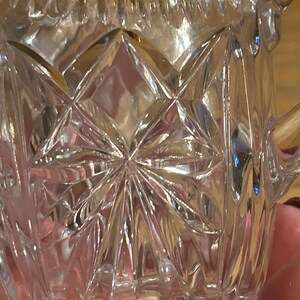 Op de afbeelding: Een glazen roomkan met een stervormig patroon. De roomkan heeft een handvat en een tuit.