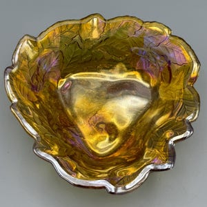 May include: An iridescent gold-colored glass bowl with a leaf design. The bowl has a scalloped edge with a silver rim. The interior of the bowl has a shiny, reflective surface. The bowl is likely vintage or antique.