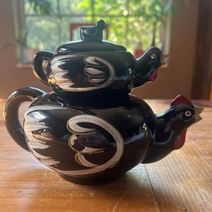 May include: Black ceramic teapot shaped like a chicken with white painted details. The teapot has a smaller teapot lid shaped like a chicken on top.