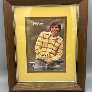 May include: A rectangular wooden picture frame with a gold inner frame and a yellow mat. The frame holds a photo of a person wearing a yellow and red sweater. The text "Empress" is visible.