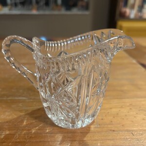Op de afbeelding: Een heldere glazen roomkan met een geslepen glasontwerp. De roomkan heeft een handvat en een tuit.