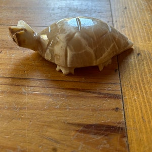 May include: A small, carved turtle figurine made of light brown and white marble. The turtle is in a walking position with its head raised.