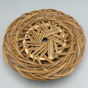 May include: A round, woven rattan basket with a central starburst design. The basket is made of thin, light brown strips of rattan, intricately woven in a circular pattern. The open weave allows for air circulation and adds to its rustic charm.
