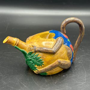 May include: A brown ceramic teapot shaped like a monkey sitting in a yellow banana. The monkey is painted with blue, green, and red details. The teapot has a brown handle and a bamboo spout.