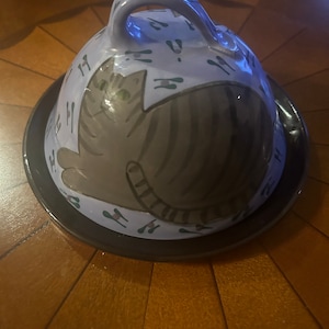 May include: A purple ceramic dome with a cat design and a handle. The dome has a black base and is decorated with a repeating pattern of paw prints.