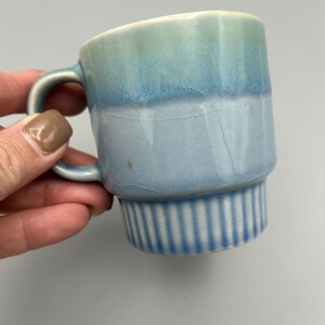 May include: A small, ceramic mug with a handle. The mug features a gradient of colors, transitioning from a light green at the top to a light blue at the bottom. The base has vertical ribbed detailing.