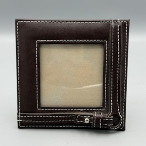 May include: A square dark brown photo frame with white contrast stitching along the edges. The frame features a central square opening and a decorative horizontal strap across the bottom with a small silver-toned metal stud detail.