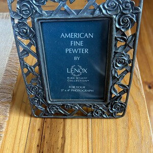 May include: A silver picture frame with a rose design. It says "American Fine Pewter by Lenox Kirk Stieff Collection" and "For Your 7.6 cm x 10.2 cm Photograph".