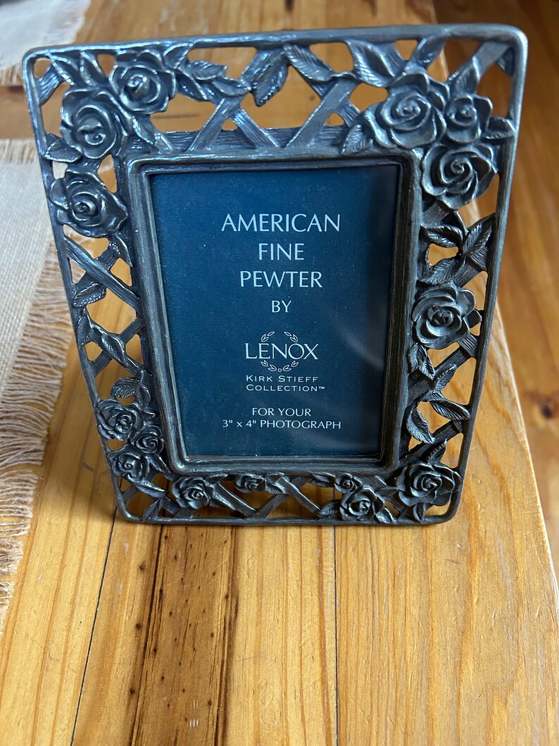 May include: A silver picture frame with a rose design. It says "American Fine Pewter by Lenox Kirk Stieff Collection" and "For Your 7.6 cm x 10.2 cm Photograph".