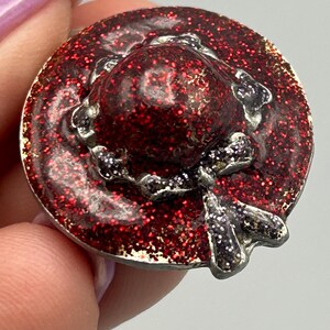 May include: A red glitter hat pin with a silver trim and a bow. The hat is a classic style with a wide brim and a small crown.