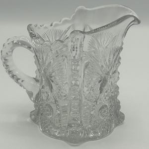 May include: A clear glass creamer with a cut glass design. The creamer has a handle and a flared lip.