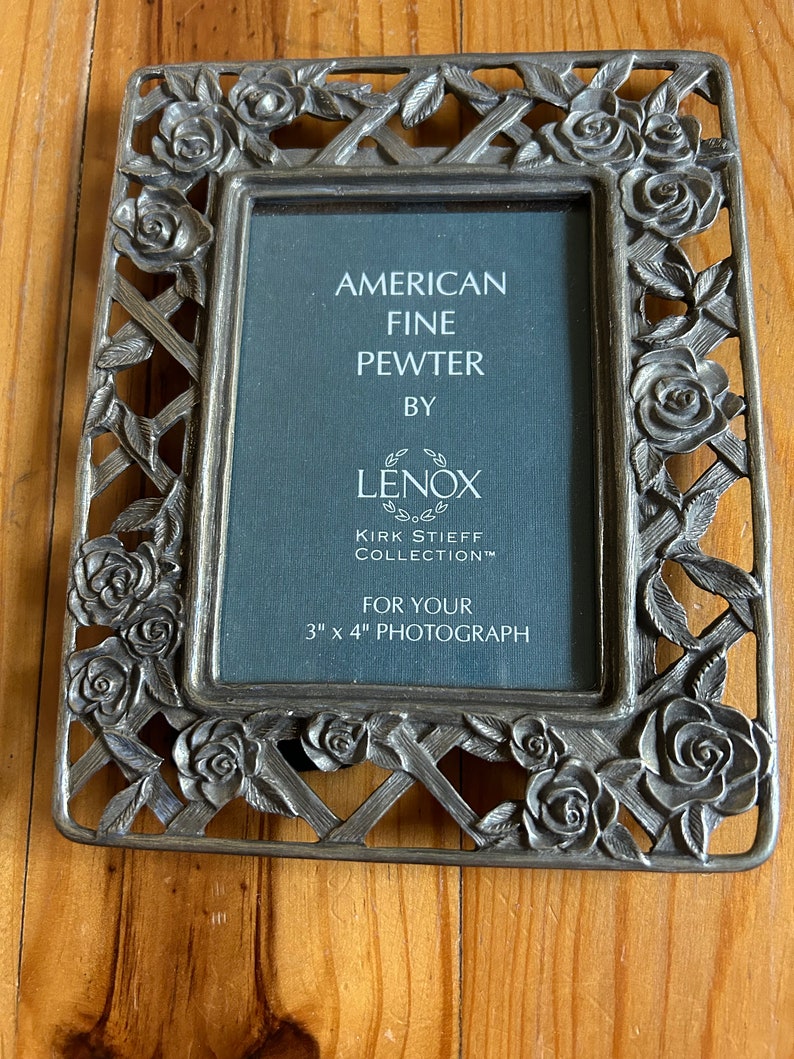 May include: A silver-toned pewter picture frame with a rose design. The frame is for a 3 x 4 inch photograph. It says "American Fine Pewter by Lenox Kirk Stieff Collection".