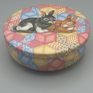 May include: A round metal tin featuring a patchwork quilt pattern in shades of pink, yellow, and blue. The lid displays two rabbits, one black and white and one brown, resting on the quilted surface. The container has a matching patterned base.