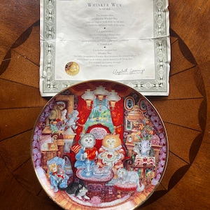 May include: A porcelain collector plate featuring a whimsical illustration of cats enjoying tea time in a cozy setting. The plate is decorated with gold trim and features the text "Whisker Wuv" and "The Franklin Mint".