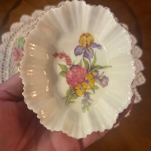 May include: A small, white, scalloped porcelain dish with a gold rim. The dish features a floral design with pink, yellow, blue, and purple flowers.