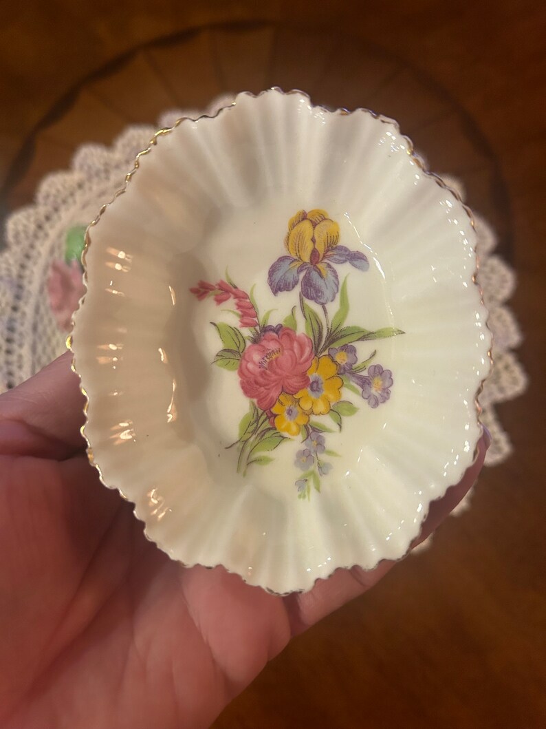 May include: A small, white, scalloped porcelain dish with a gold rim. The dish features a floral design with pink, yellow, blue, and purple flowers.