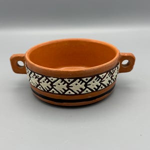 May include: A small, round, terracotta-colored bowl with two handles. The rim features a decorative band with a repeating pattern of white and black geometric shapes. Two black stripes encircle the bowl near the base.