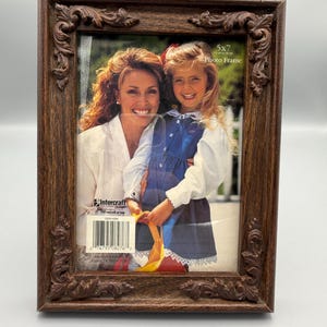 May include: A rectangular dark brown photo frame with ornate carved scrollwork detailing along the edges. The frame holds a display insert featuring a photograph of an adult and a child, with text reading 5x7 and Intercraft printed on the front.