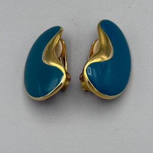 May include: A pair of clip-on earrings with a teal enamel finish and gold-tone trim. The earrings have a teardrop shape with a curved design. The clip-on backs are visible. The earrings are displayed on a light gray surface.