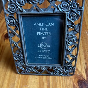 May include: A silver-toned pewter picture frame with a rose design. The frame is for a 3x4 inch photograph and says "American Fine Pewter by Lenox Kirk Stieff Collection".