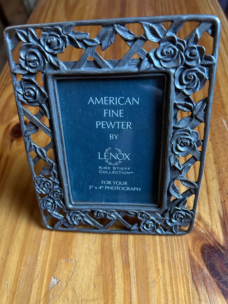 May include: A silver-toned pewter picture frame with a rose design. The frame is for a 3x4 inch photograph and says "American Fine Pewter by Lenox Kirk Stieff Collection".