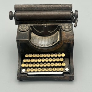 May include: A vintage-style metal typewriter pencil sharpener with a black and silver finish. The sharpener has a small, round, black handle on the right side and a larger, round, black handle on the left side. The sharpener has a metal roller on top and a keyboard with the letters QWERTYUIOPASDFGHJKLZXCVBNM and numbers 1234567890.