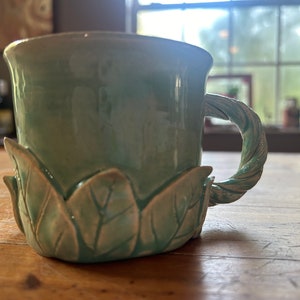 May include: A light green ceramic mug with a twisted handle and a base sculpted with leaves.