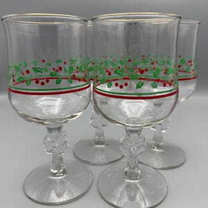 May include: Set of four clear wine glasses with gold rims. Each glass features a festive design of green holly leaves and red berries, with a red and green stripe near the base. The stems have a decorative floral design.