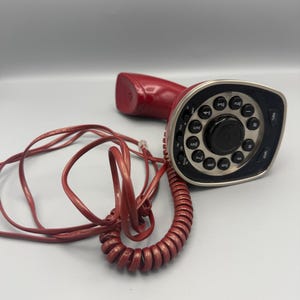 May include: A red corded telephone featuring a circular keypad with black buttons and white numbers. The device has a silver-toned trim around the dial area, a matching red handset, and a coiled red telephone cord extending from the base unit.