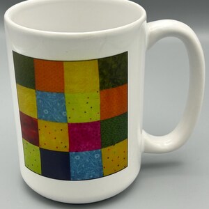 May include: White ceramic mug with a colorful patchwork design. The design features squares of various colors, including red, orange, yellow, green, blue, and pink.