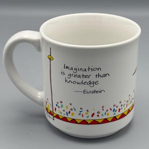 May include: White ceramic mug with a handle, featuring the quote "Imagination is greater than knowledge" by Einstein. The mug has colorful decorative accents, including a red and yellow border with a pattern of triangles and dots.