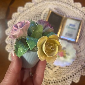 May include: A porcelain figurine of a bouquet of flowers in a blue pot. The bouquet includes a yellow rose, pink carnations, and green leaves.