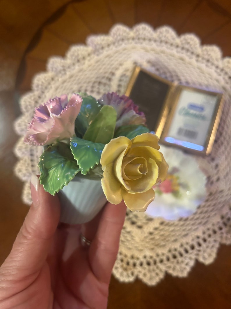 May include: A porcelain figurine of a bouquet of flowers in a blue pot. The bouquet includes a yellow rose, pink carnations, and green leaves.