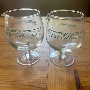 May include: Two clear glass goblets with a green design and the text "Pat O'Brien's" on the front. The text "Have Fun!" is written on the side of each goblet.