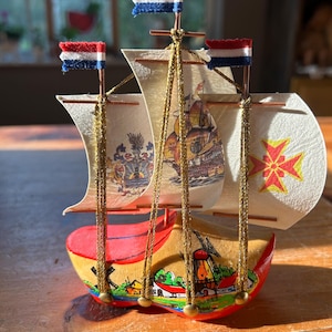 May include: A miniature wooden ship with sails, a red, white, and blue flag, and a wooden clog base. The ship is decorated with a Dutch windmill and other Dutch-themed imagery.