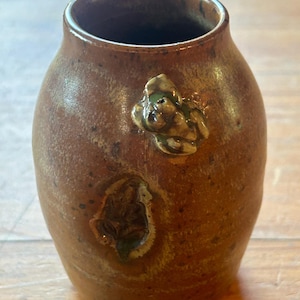 May include: A small, brown ceramic vase with a textured surface and a green and brown floral design. The vase has a small, round opening at the top.