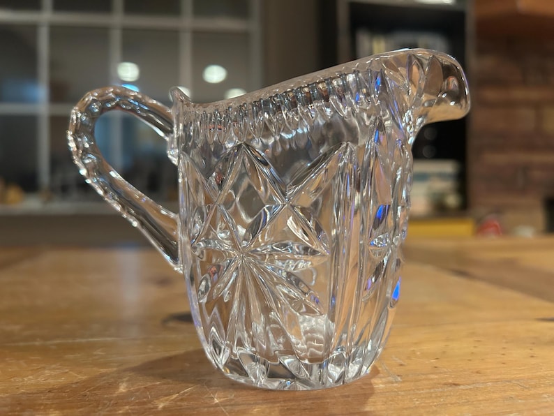 Op de afbeelding: Een heldere glazen roomkan met een geslepen glasontwerp. De roomkan heeft een handvat en een tuit.