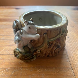 May include: A ceramic planter shaped like a tree trunk with a squirrel figurine climbing on it. The planter is brown and green with a white interior.