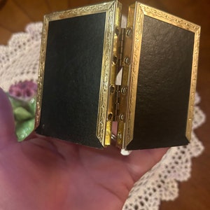 May include: A gold-colored, hinged picture frame with a black velvet interior. The frame has a decorative, ornate design.