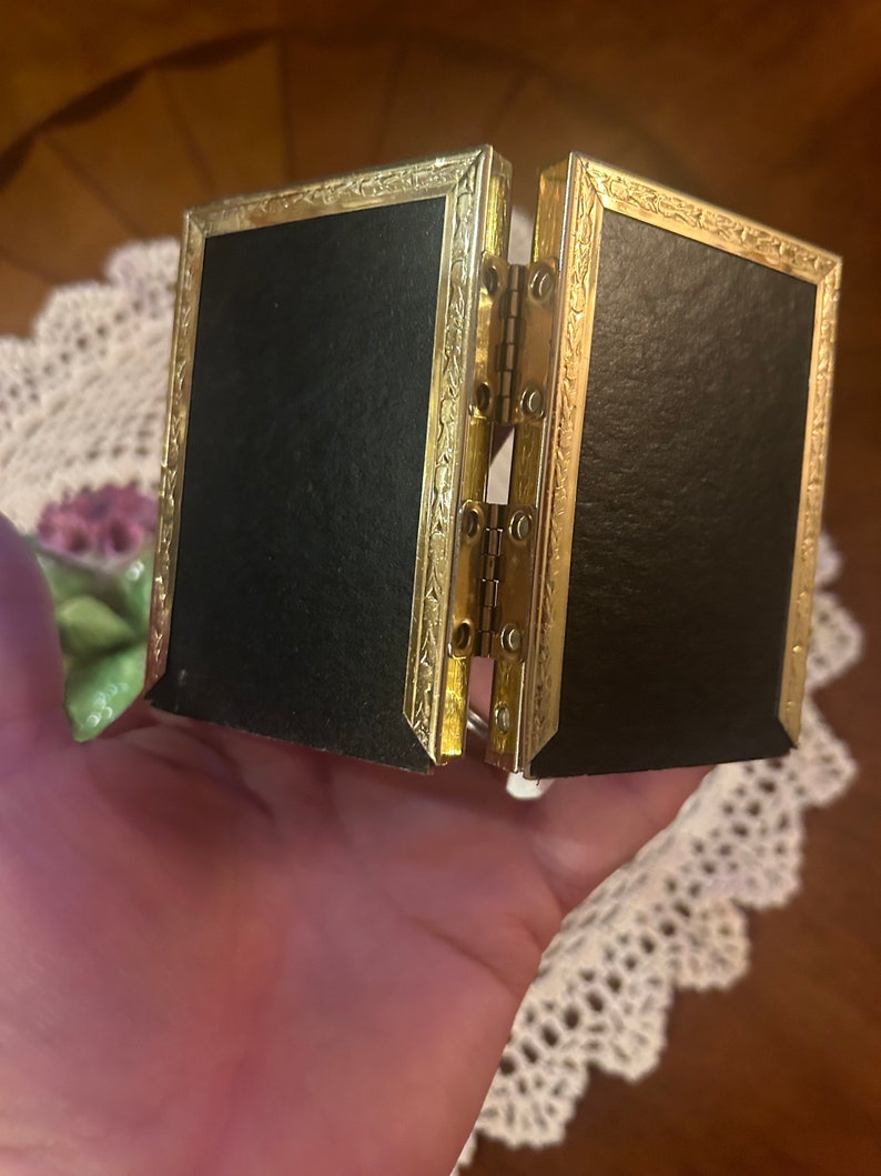 May include: A gold-colored, hinged picture frame with a black velvet interior. The frame has a decorative, ornate design.