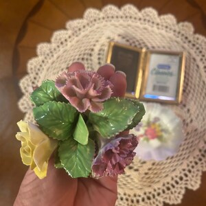 May include: A hand holding a porcelain flower bouquet with yellow, pink, and green flowers. The bouquet is made of delicate porcelain and features realistic details.