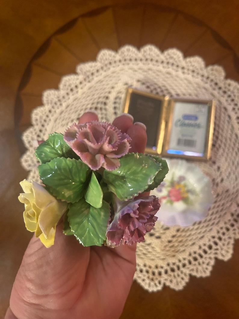 May include: A hand holding a porcelain flower bouquet with yellow, pink, and green flowers. The bouquet is made of delicate porcelain and features realistic details.