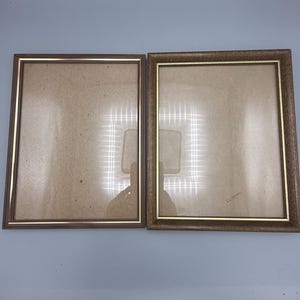 May include: Two rectangular picture frames with brown borders and thin gold inner trim. The left frame has a smooth finish, while the right frame features a textured, wood-grain appearance. Both frames contain clear glass panels.