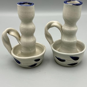 May include: Two handmade ceramic candle holders. The candle holders are off-white with a blue rim and blue leaf-shaped designs. Each holder has a handle and a circular base to catch wax. The candle holders are a unique home decor item.